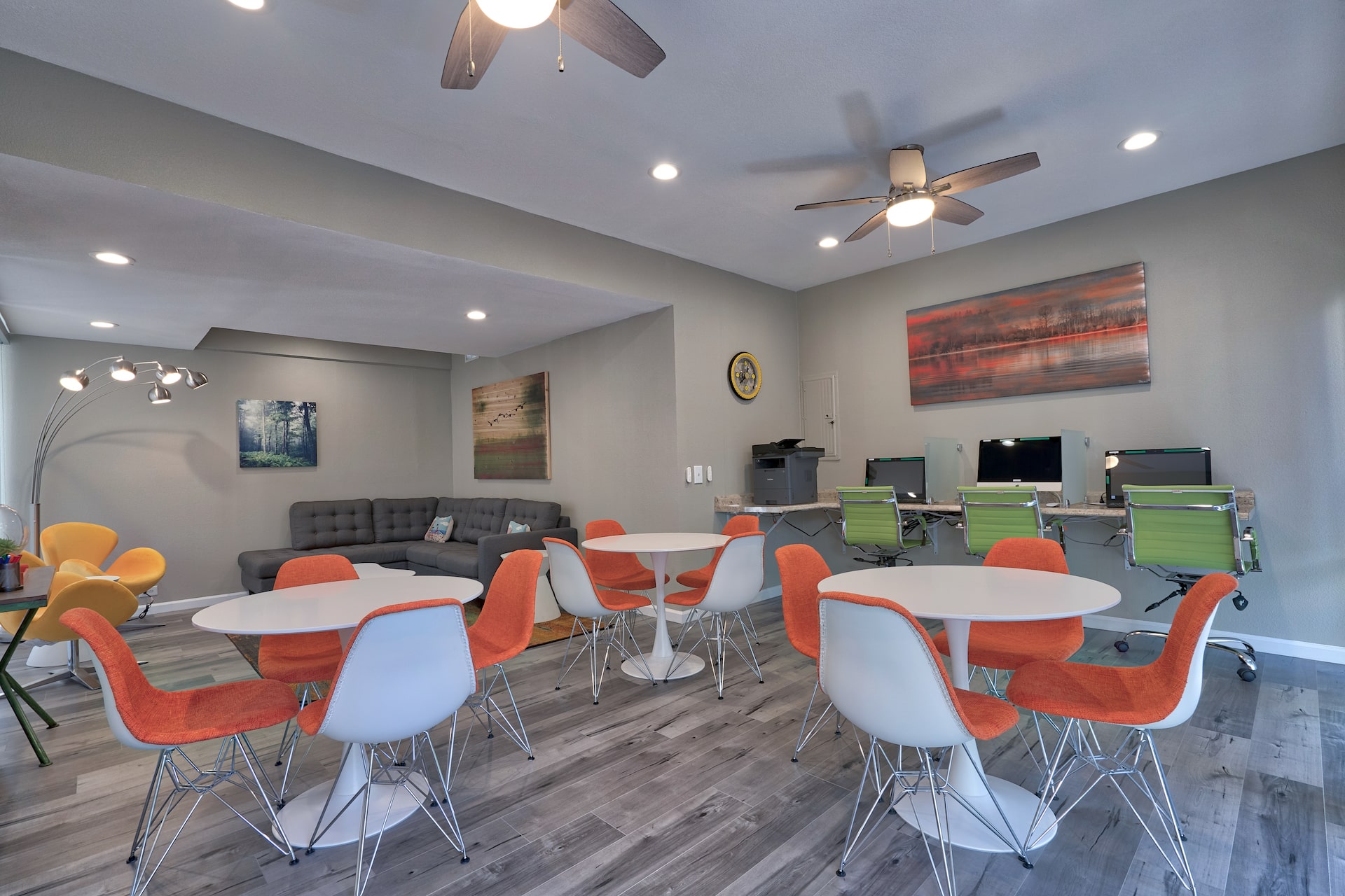 common area lounge with modern circular wine-stem tables, a row of computers along the wall, and a large L shaped couch in the corner.