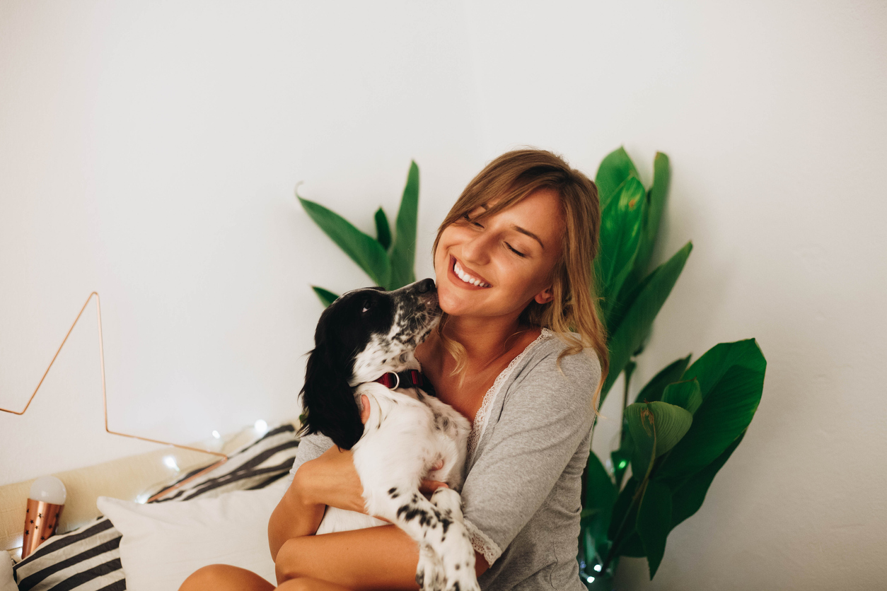 Young adult sitting in bed holding a dog while it licks their face.